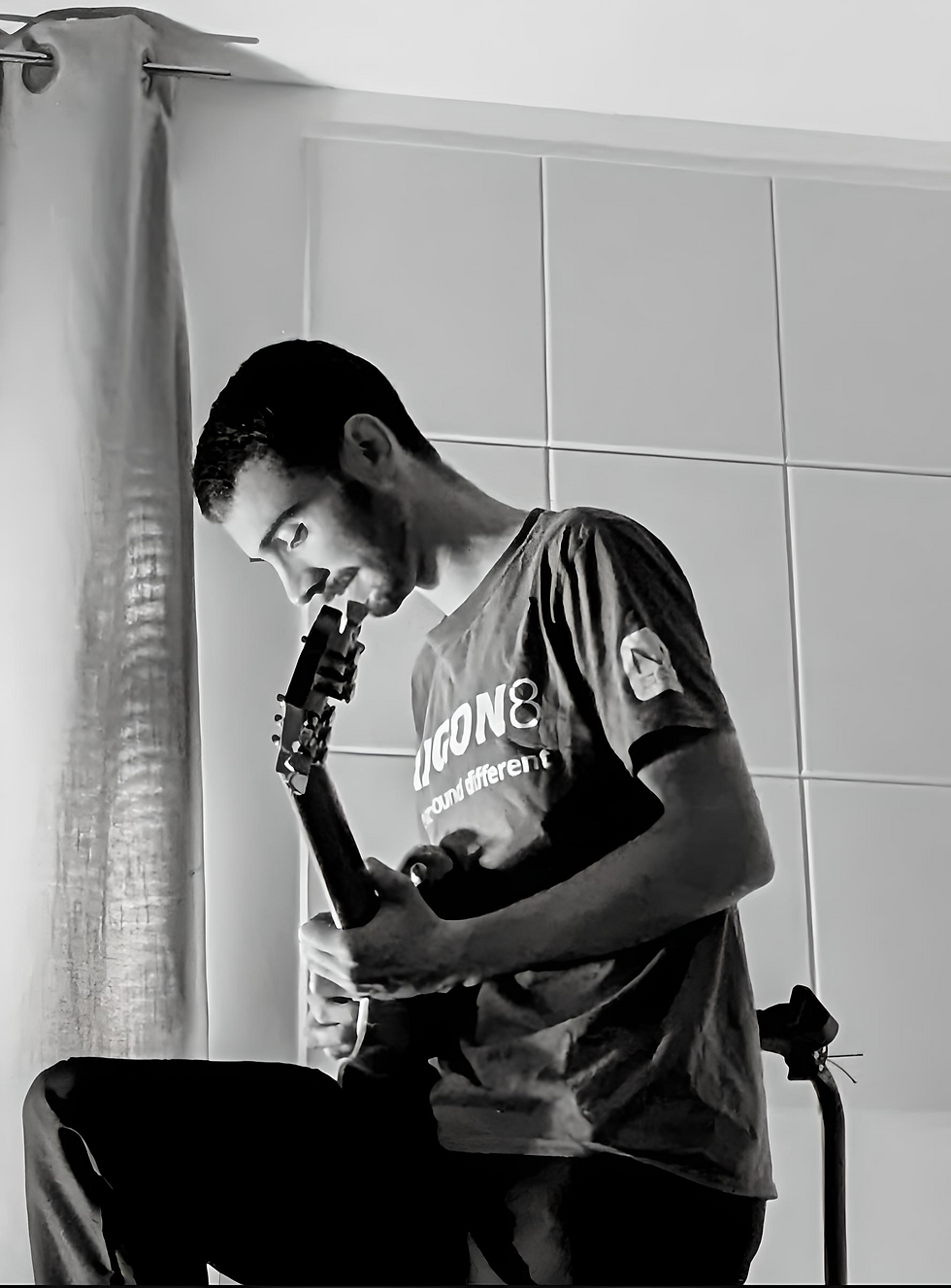 Man playing guitar in a room with tiled walls, soft lighting. Wearing a logo T-shirt, looking down focused. Curtain partially visible. Zachary Mason - The Cage, a music blog powered by Cage Riot