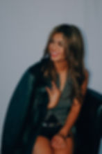 Smiling woman with long hair wearing a leather jacket and dark outfit, seated against a plain background, exuding a cheerful mood. Ashley Mora - The Cage, a music blog powered by Cage Riot