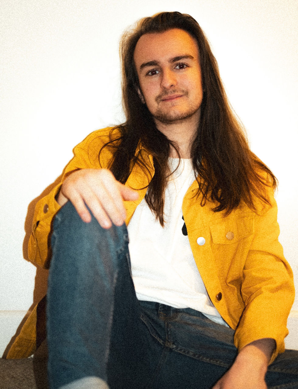 Person with long hair in a yellow jacket and jeans sits casually against a white wall, smiling slightly, creating a relaxed atmosphere. Rhys Hurd - The Cage, a music blog powered by Cage Riot