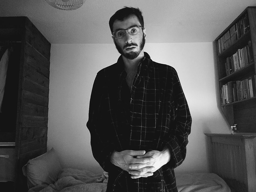 Man in glasses wearing a plaid robe stands in a dimly lit room with a bookshelf and bed. Mood is calm and introspective. Zachary Mason - The Cage, a music blog powered by Cage Riot
