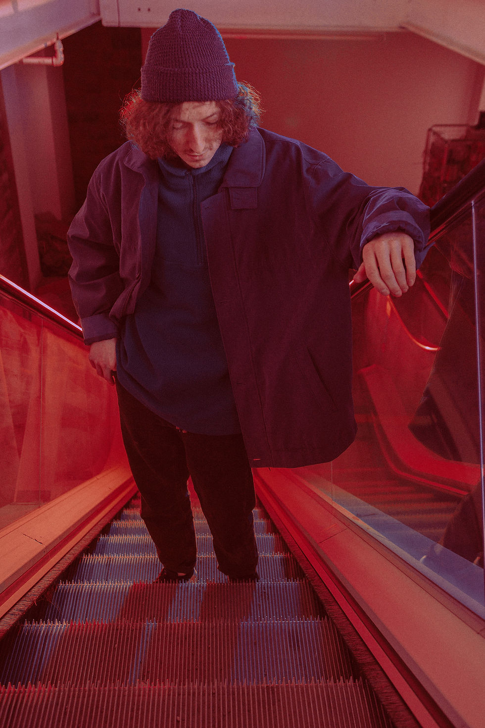 Person in dark clothing and a beanie descends an escalator. Red lighting creates a moody atmosphere, with reflective glass sides. In VideColour - The Cage, a music blog powered by Cage Riot