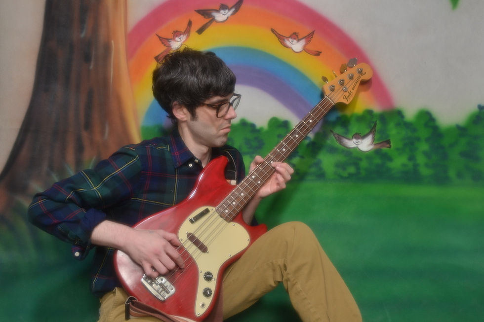 Man playing a red guitar in front of a colorful mural with a rainbow and birds. Wears glasses and checkered shirt, focused expression. Make Believe Love - The Cage, a music blog powered by Cage Riot