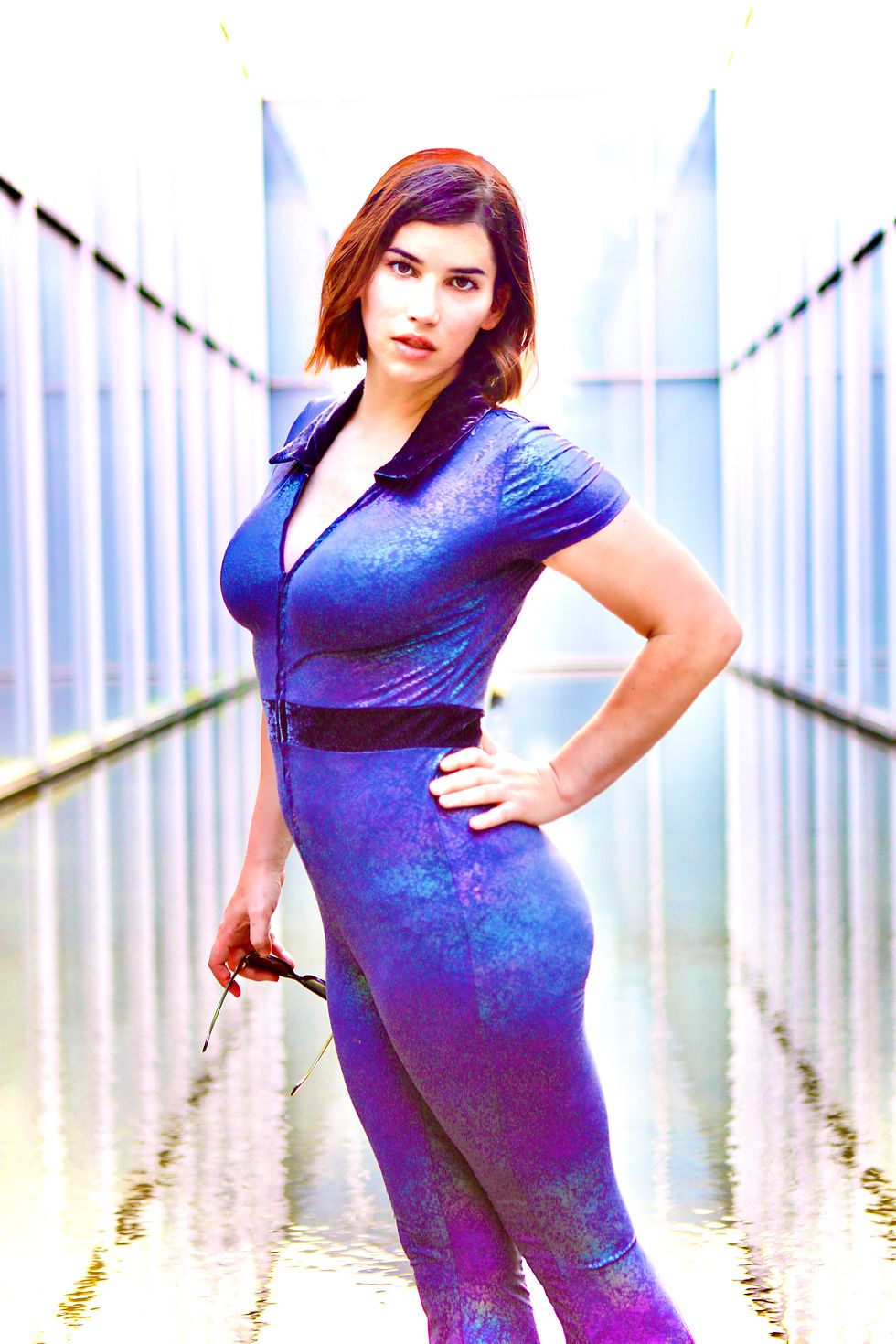 Woman in a purple jumpsuit stands confidently in a modern corridor with water reflections, holding sunglasses, against a bright background. SERAh - The Cage, a music blog powered by Cage Riot