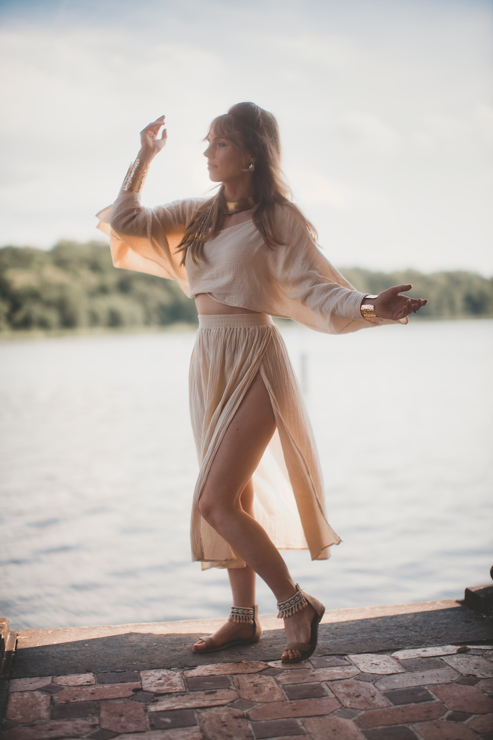 A woman in a beige outfit poses gracefully by a lake, with sunlight casting shadows. She's wearing a flowy skirt and sandals. Marie Chain - The Cage, a music blog powered by Cage Riot