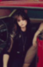 Woman with long hair sits in a vintage red car, looking pensive. She's wearing a lace top and a necklace, with a retro interior visible. Emma Forgette - The Cage, a music blog powered by Cage Riot