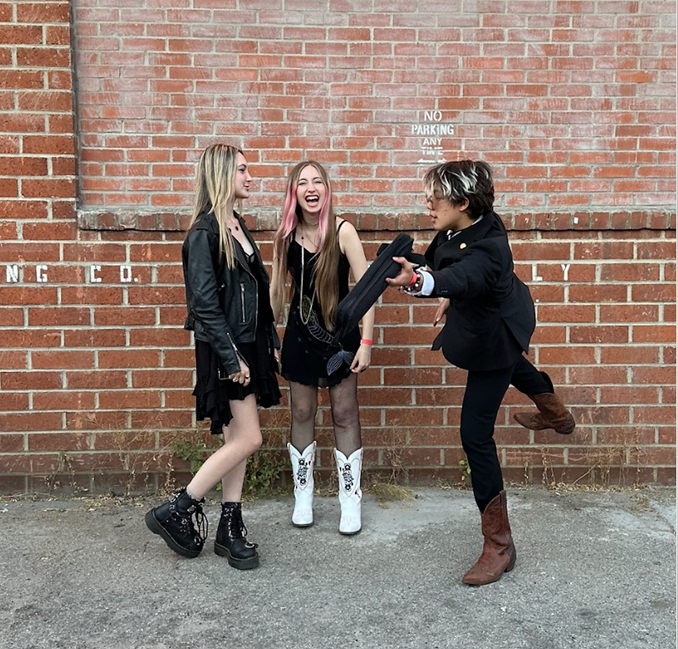 Three people in black outfits laughing by a brick wall with "No Parking" text. One person playfully kicks in cowboy boots. Casual mood. LED - The Cage, a music blog powered by Cage Riot