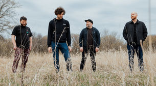 Four men stand in a grassy field under a cloudy sky, wearing casual clothes. One looks down, others smile, conveying a relaxed mood. The Winter Sloths - The Cage, a music blog powered by Cage Riot