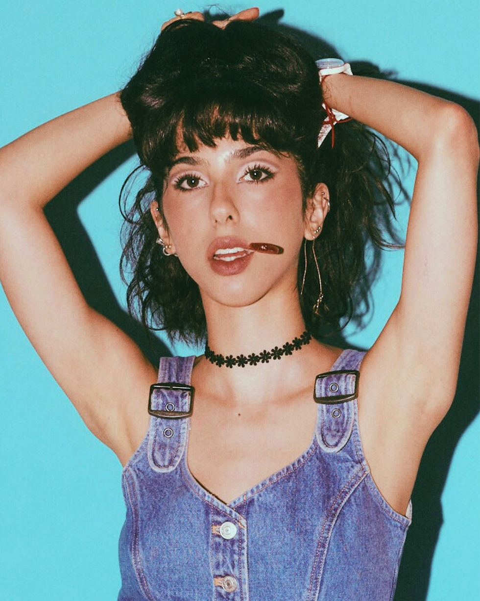 Woman in denim outfit with arms up, holding a lollipop in her mouth. She stands against a bright blue background, conveying a playful mood. Neska Rose’ - The Cage, a music blog powered by Cage Riot