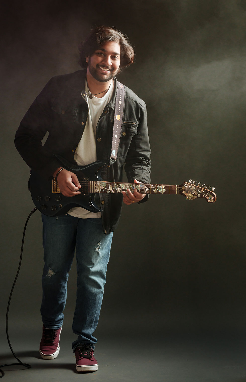 Man playing electric guitar, wearing a black jacket, ripped jeans, and red sneakers. Dark smoky background, smiling expression. Varun Sheel - The Cage, a music blog powered by Cage Riot