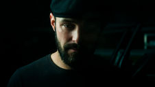 A man with a beard and cap gazes downward in low light. Dark background; mood is contemplative. No visible text or notable colors. Seth Schaeffer - The Cage, a music blog powered by Cage Riot