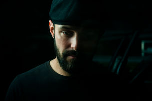 A man with a beard and cap gazes downward in low light. Dark background; mood is contemplative. No visible text or notable colors. Seth Schaeffer - The Cage, a music blog powered by Cage Riot