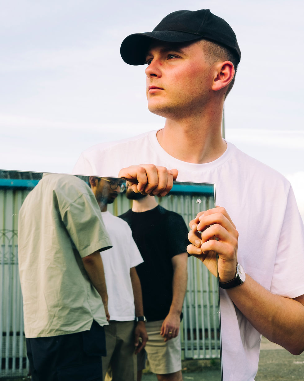 Man in black cap holds a mirror reflecting two people. Wearing white shirt, outdoors setting, bright sky. Calm and thoughtful mood. SPEAK! - The Cage, a music blog powered by Cage Riot
