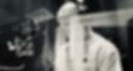 Black and white image of a person in a studio, wearing headphones, standing behind a mesh screen, with a microphone in front, creating a focused mood. Ben Heyworth - The Cage, a music blog powered by Cage Riot