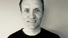 Man in a black t-shirt smiles slightly against a plain background. Image is in black and white. Calm and neutral expression. Formoe - The Cage, a music blog powered by Cage Riot