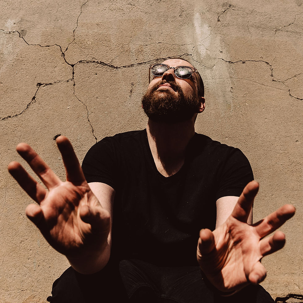 Bearded man in sunglasses extends open hands towards the camera. He's sitting against a cracked beige wall, bathed in warm sunlight. Rhys Tranter - The Cage, a music blog powered by Cage Riot