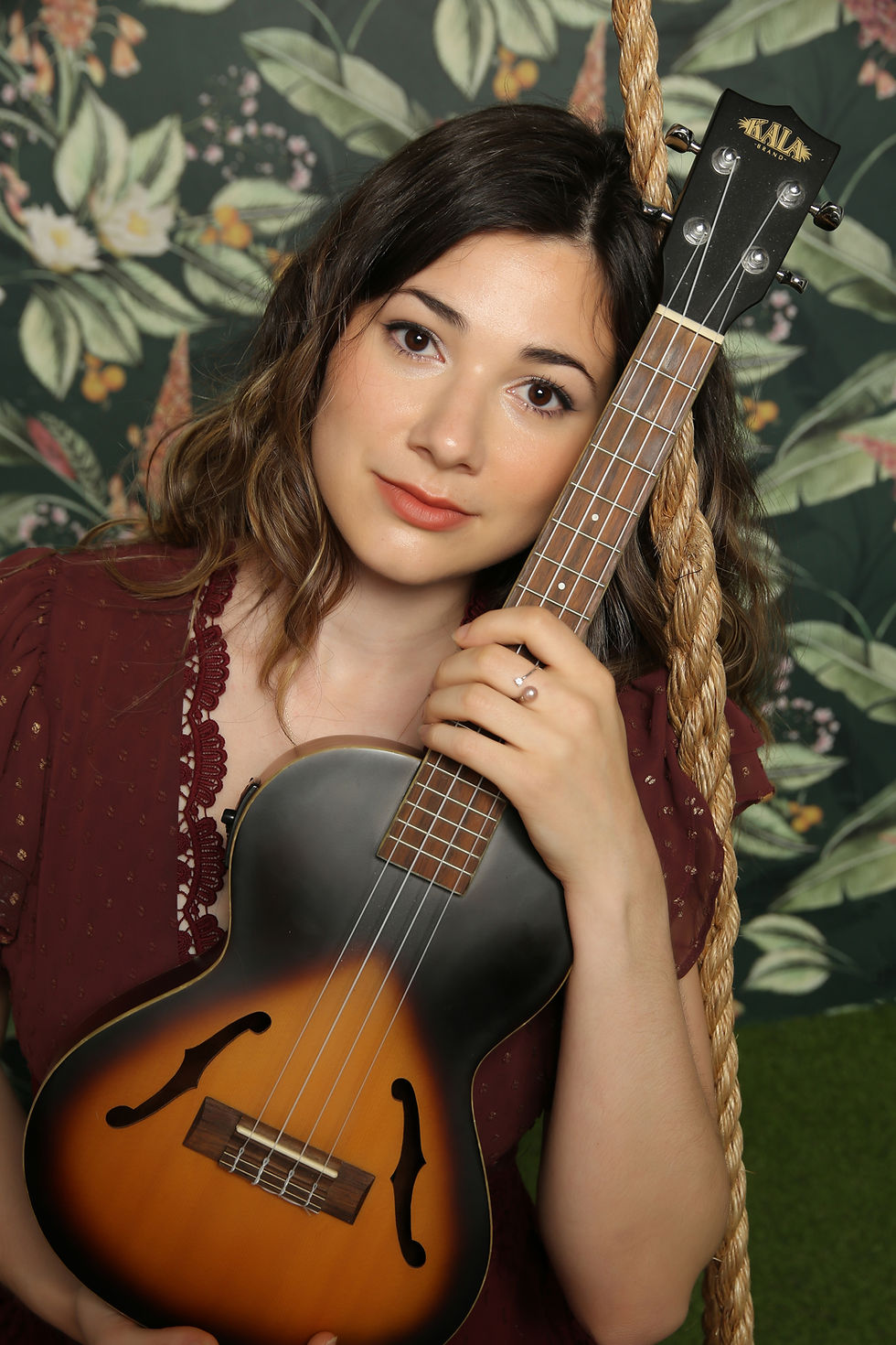 Woman in maroon dress holds ukulele, leaning on rope swing. Floral wallpaper background. Calm expression. Brand "Kala" visible on instrument. Talon David - The Cage, a music blog powered by Cage Riot
