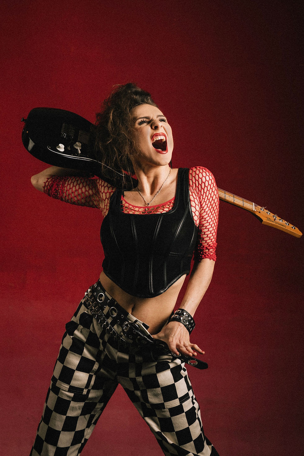 Woman in fishnet top and checkered pants holds guitar, shouting passionately. Red background, intense expression, rock vibe. Electric Lady - The Cage, a music blog powered by Cage Riot