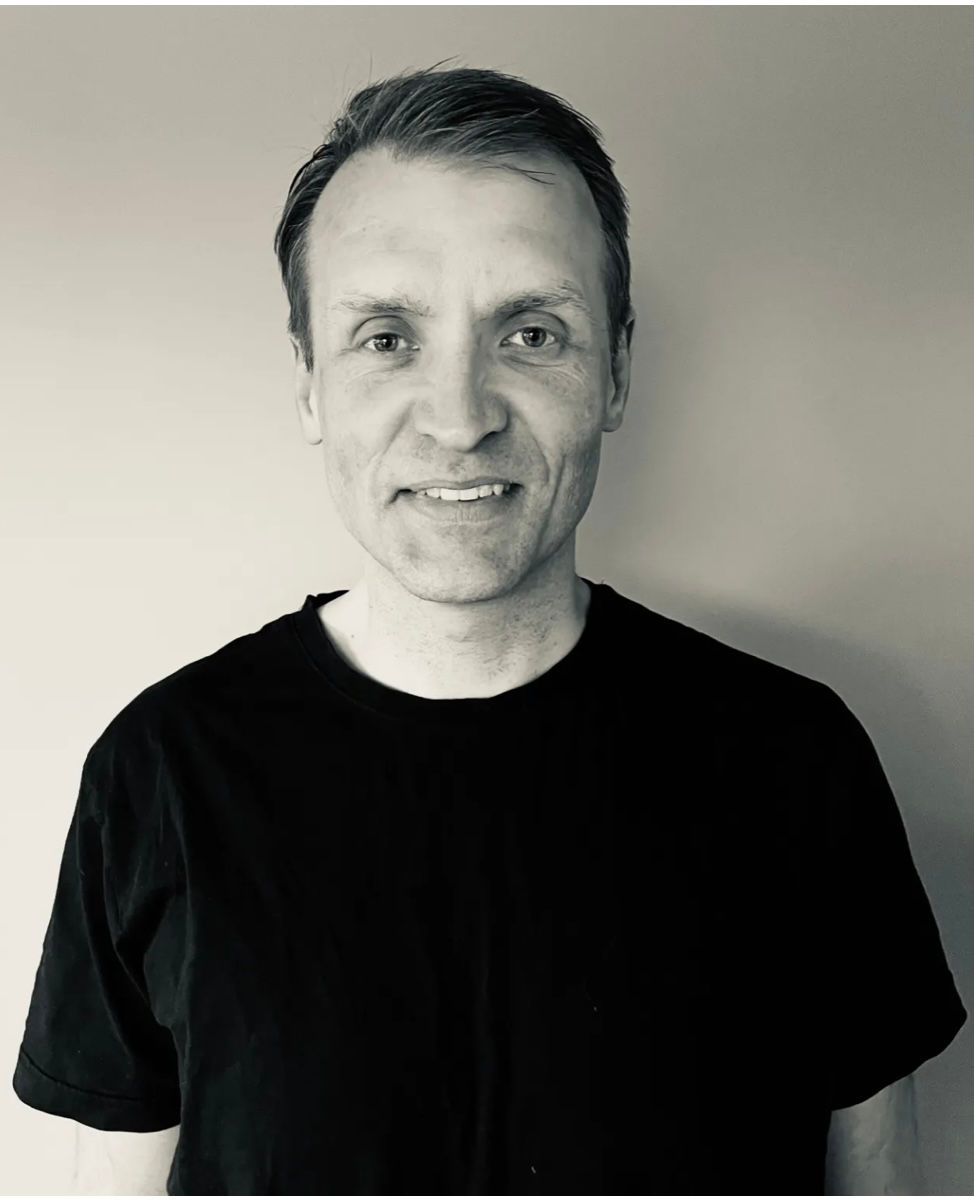Man in a black t-shirt smiles slightly against a plain background. Image is in black and white. Calm and neutral expression. Formoe - The Cage, a music blog powered by Cage Riot