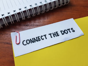 Paper note clipped to a yellow folder reads CONNECT THE DOTS beside a spiral notebook on a wooden desk.