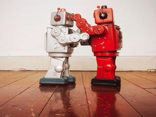 Two toy robots, one silver and one red, face each other as if boxing on a wooden floor. The background is a plain white wall.