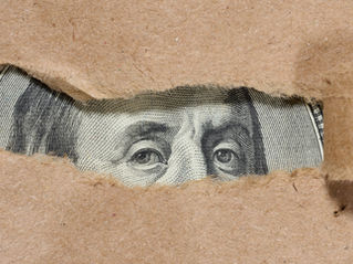 Torn brown paper reveals part of a U.S. bill with eyes in focus, creating a sense of intrigue. The background is light brown.