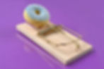 Sugar-coated donut on a mousetrap against a purple background, creating a playful but risky still life.