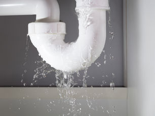 Leaking white PVC pipe joint spraying water indoors against a gray wall, with splashes and droplets everywhere