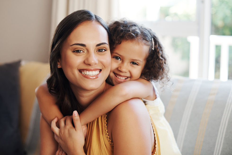 Smiling woman and child embrace on a sofa. The child is hugging her from behind. Bright, cozy room with striped cushions.