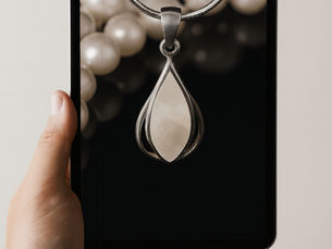 Hand holding a tablet displaying a silver pendant with a teardrop shape on a dark background. Pearls are visible in the background.