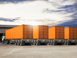 Orange shipping containers on trailers lined up in an industrial area. Overcast sky and warehouses in the background.