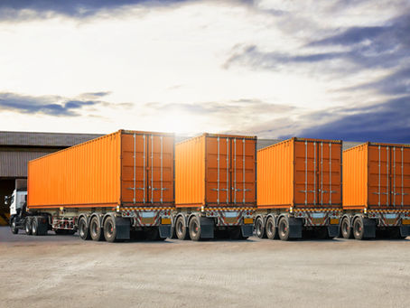 Orange shipping containers on trailers lined up in an industrial area. Overcast sky and warehouses in the background.
