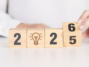 Hands are changing wooden blocks from 2025 to 2026 on a white surface. A lightbulb icon is engraved on the second block.