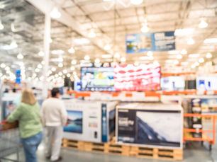 Blurry image of two people shopping in an electronics store with numerous TVs displayed. Bright lighting and banners in the background.