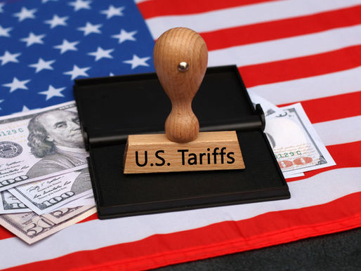 Wooden stamp with "U.S. Tariffs" on a black ink pad, U.S. flag in the background, and $100 bills visible, conveying economic theme.