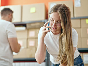 Woman with glasses talks on a phone in a warehouse, with stacked boxes and a blurred man in the background.