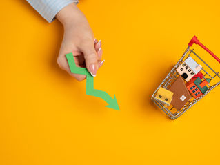Hand holds a green downward arrow beside a shopping cart filled with miniature houses on an orange background.