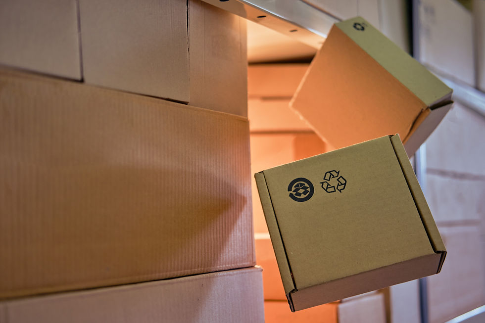 Cardboard boxes in a warehouse are stacked on shelves. Two boxes are falling, showing recycling symbols. Warm lighting fills the space.