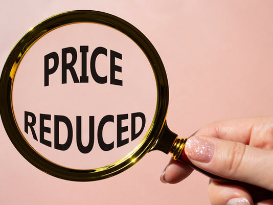 Hand holding magnifying glass over "PRICE REDUCED" text on pink background. The magnifying glass has a golden rim.