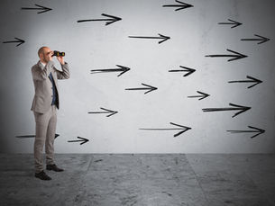 Businessman in a gray suit looks through binoculars at a wall covered with black arrows in a sparse concrete room.