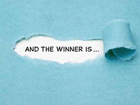 Torn blue paper revealing text "AND THE WINNER IS..." on white background. The paper curl creates suspense and excitement.