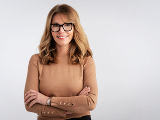 Woman in brown sweater and glasses smiling with arms crossed. Neutral white background, relaxed and confident expression, casual setting.