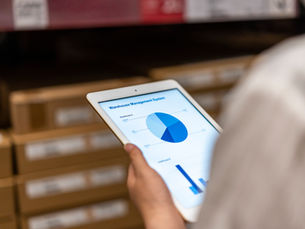 Person in a warehouse holds a tablet showing a Warehouse Management System dashboard with blue pie and bar charts.