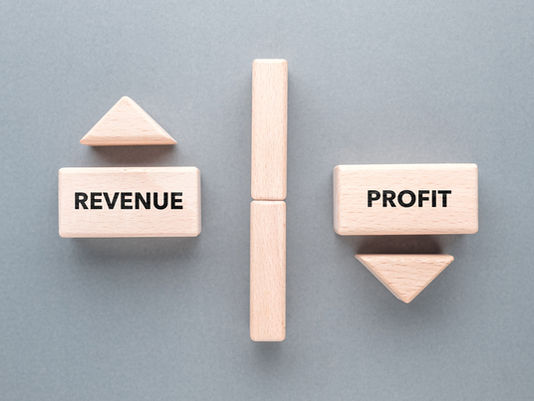 Wooden blocks on a gray background. "REVENUE" block has an upward arrow, "PROFIT" block has a downward arrow, separated by vertical blocks.