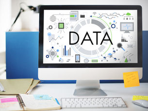 Computer screen with the word "DATA" surrounded by charts and icons, on a desk with books, notebooks, and sticky notes. Office setting.