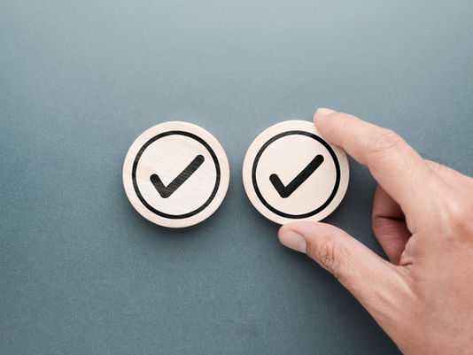Hand holding a wooden disk with a checkmark, next to another similar disk, on a gray background. Symbolizes approval or completion.