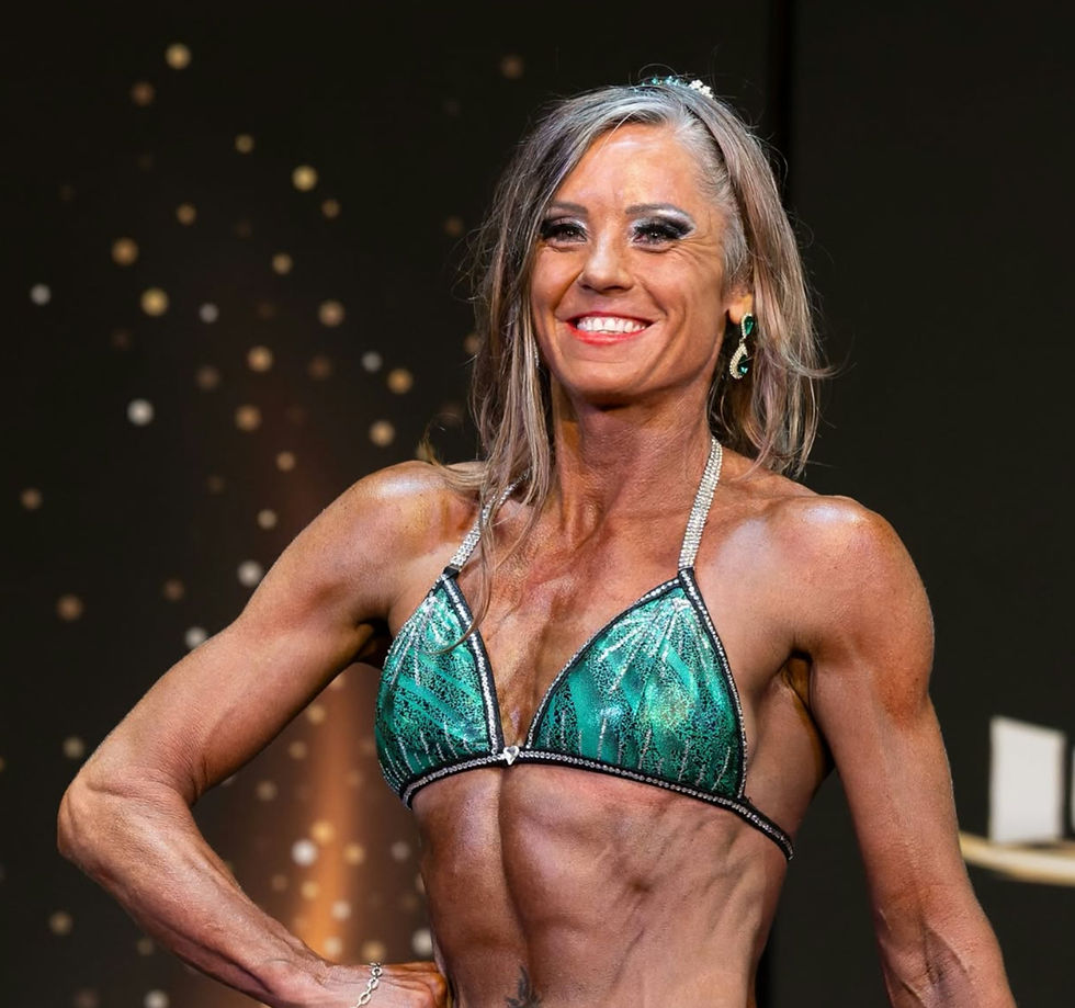 Smiling female bodybuilder in a green bikini poses onstage against a dark, sparkly backdrop, looking confident and proud.