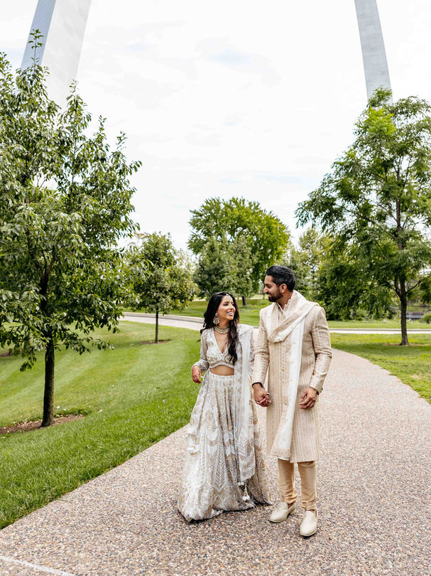 Smiling couple walk down a path in a park in front of the St. Louis, MO Gateway Arch in the background.