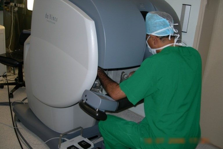A surgeon in green scrubs operates the da Vinci robotic system in a hospital setting. The room appears clean and sterile.