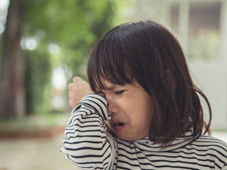 Young child in a striped shirt crying, wiping tears with their arm. Background shows a blurred outdoor setting with green trees. Joyful Seeds Paediatric and Developmental Clinic located in Bukit Timah.
