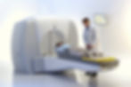 A doctor assists a patient into an MRI machine in a sterile room. The machine is white and gray, and a monitor displays graphs on the wall.